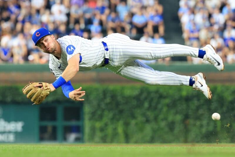 A baseball player in a blue and white pinstriped uniform dives fully extended in midair, attempting to catch a ball just above the grass, with a blurred crowd in the background.