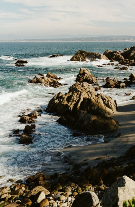 Rocky coastline with waves crashing against large stones, foamy surf, and a distant view of the ocean and hazy mountains under a partly cloudy sky. Pebbles cover the beach in the foreground.