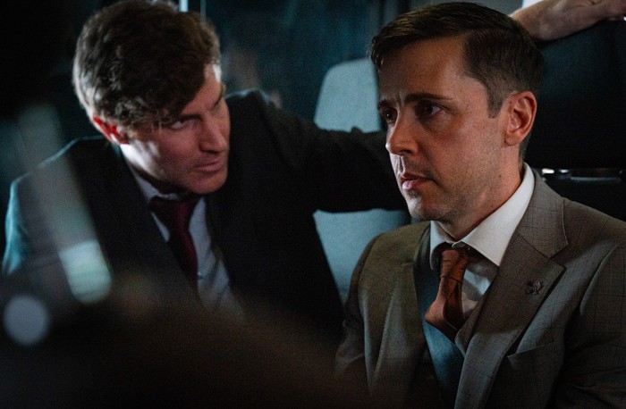 Theo looks over Todd's shoulder in a scene from Corriedale. Both men are wearing suits.
