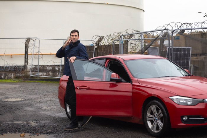 Kit stands on an industrial site next to a red car in a scene from Coronation Street