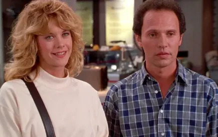 Two people standing side by side indoors. The woman has styled hair and wears a light top. The man has short hair and wears a checkered shirt