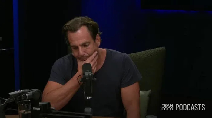 Person in a podcast studio, wearing a casual t-shirt, sitting thoughtfully with hand on chin. Microphone and "Team Coco Podcasts" logo visible