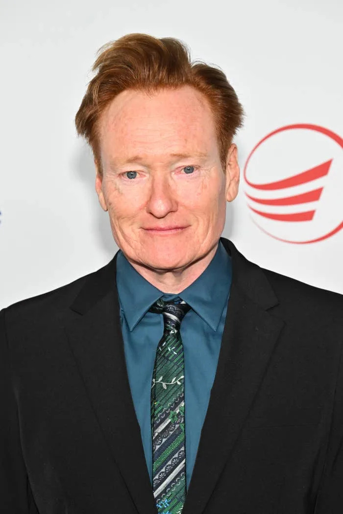 A person in a suit with a patterned tie poses on a red carpet, against a backdrop with circular design elements