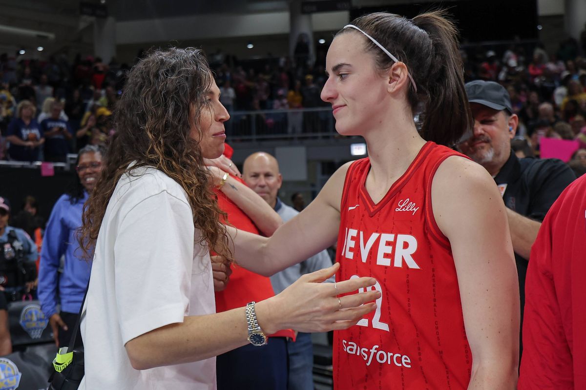 Two female individuals, one dressed in a red jersey with the word "EVER" prominently displayed, engage in a heartfelt moment, possibly post a basketball match, as they exchange words and gestures in a lively stadium setting with an audience in the background.