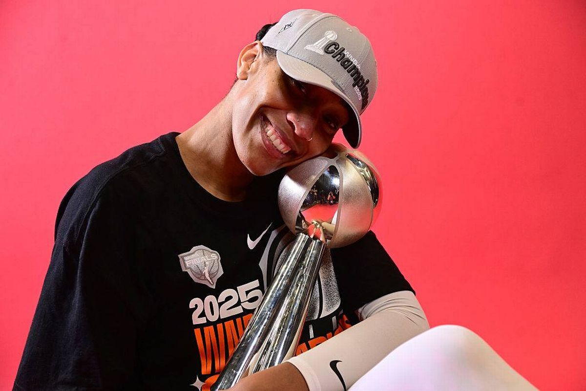 An individual wearing a cap and a black t-shirt is holding a trophy and smiling for the camera in front of a red background.