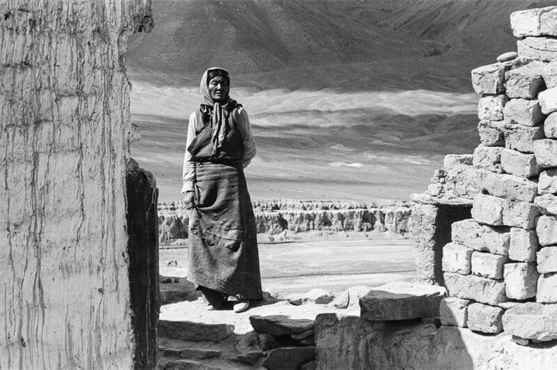 An elderly person in traditional clothing stands among old stone ruins, with mountains and a vast landscape visible in the background under natural daylight.