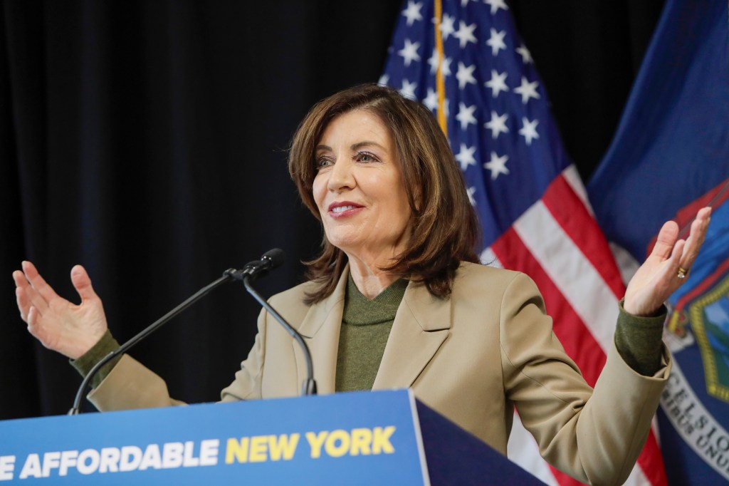 Kathy Hochul speaking at a podium with two microphones, an American flag, and a New York State flag behind her.