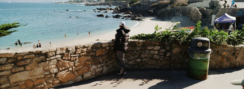 A person in a dark jacket and white cap leans on a stone wall, overlooking a beach with people swimming. A trash can is nearby, and the scene includes blue ocean water and rocky shoreline.