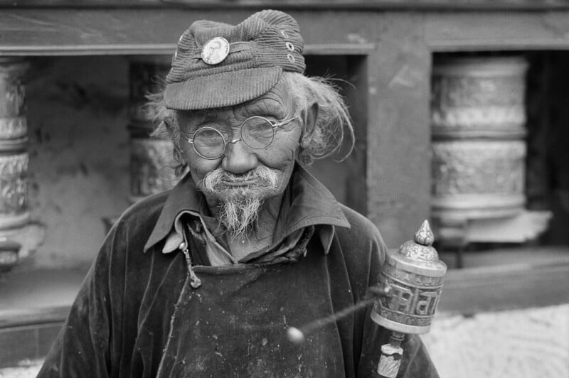 An elderly person with glasses, a cap, and a mustache holds a prayer wheel in front of large spinning prayer wheels. The image is black and white, and the person wears traditional clothing.