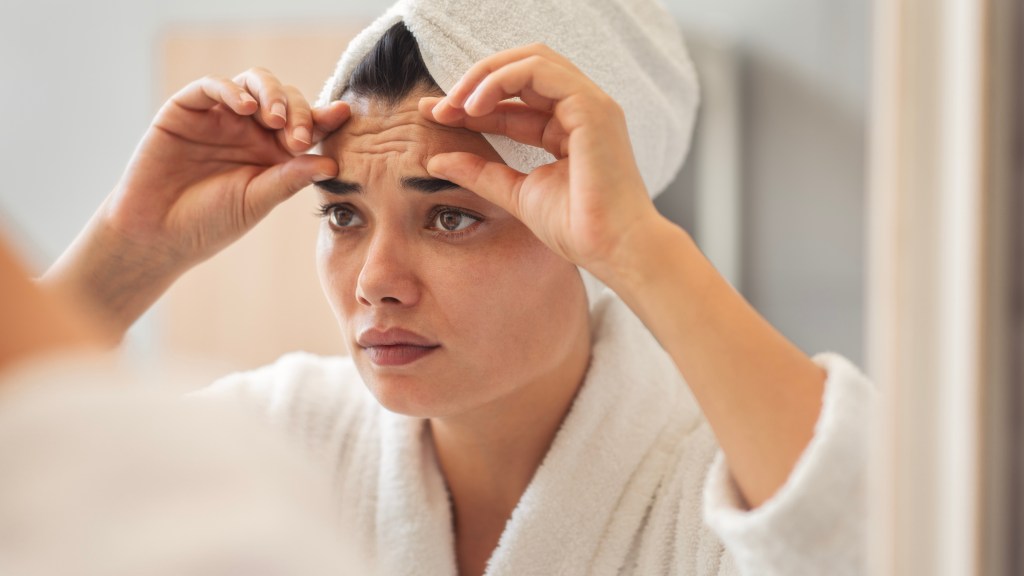 A young woman in a bathrobe and towel turban looking sadly at wrinkles on her forehead in a mirror.