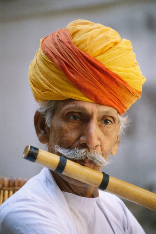 An elderly man with a white mustache and beard wears a bright yellow and orange turban. He holds a wooden flute to his lips, appearing to play, and wears a white shirt. The background is softly blurred.
