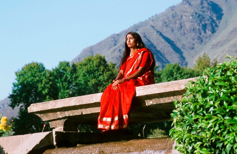 A woman in a bright red saree sits on a stone ledge outdoors, surrounded by greenery, with tree-covered hills and a clear blue sky in the background.