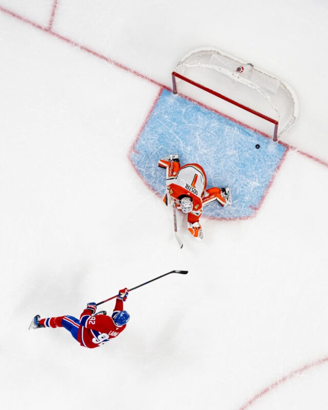 Aerial view of a hockey player in a red and blue uniform taking a shot on goal as an opposing goalie in orange and white gear prepares to block the puck on an ice rink.