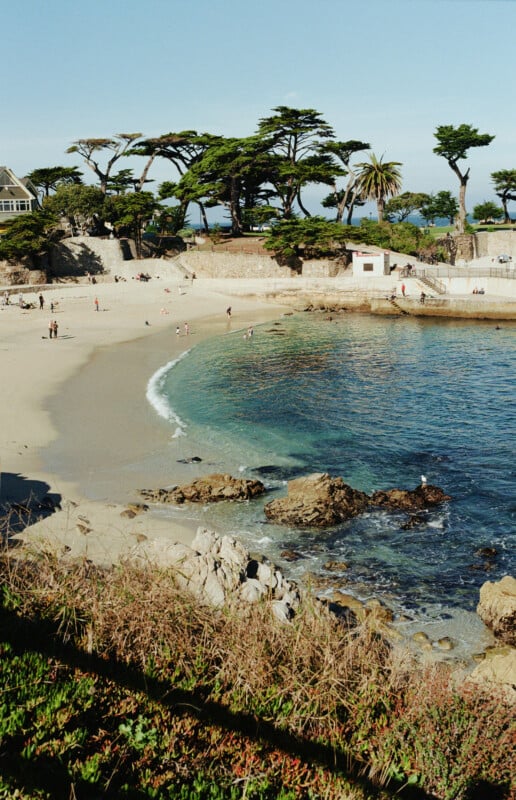 A sandy beach with clear turquoise water, rocky shoreline, and scattered people. Tall trees and a few buildings are visible in the background under a clear blue sky.
