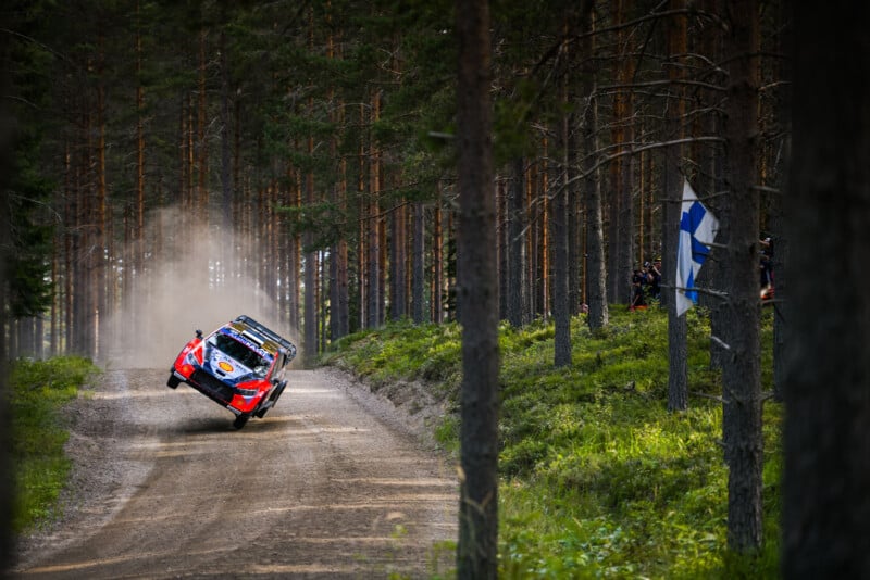 A rally car speeds through a forest dirt road, tilted on two wheels with dust trailing behind. Tall pine trees line both sides of the track, and a Finnish flag is visible on the right.
