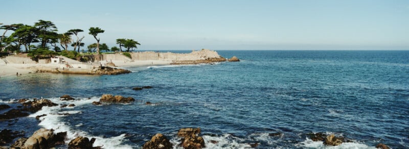 A calm coastal scene shows rocky shorelines and gentle waves, with a sandy beach and tall trees in the background under a clear blue sky.