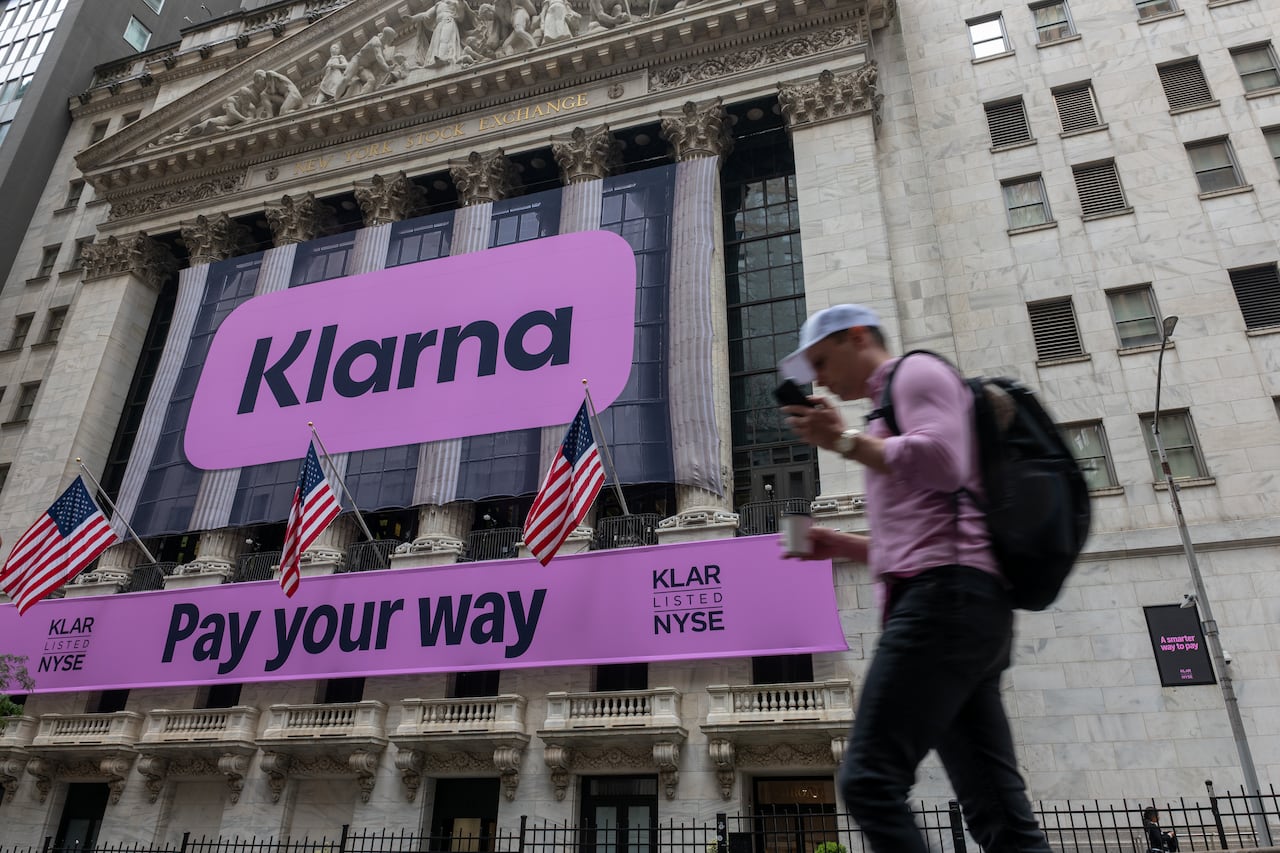 A purple banner that says 'Klarna,' and a smaller one below it that says 'Pay your way,' are hung outside a historic building. A man holding a coffee and a phone is shown in motion nearby.