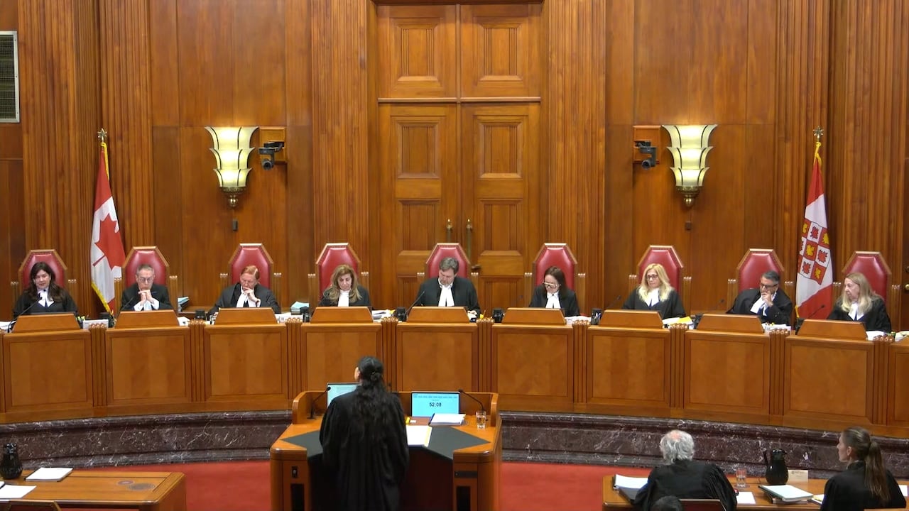 Nine people in black robes are seated in high-backed chairs in a wood-panelled room facing the camera. A woman in black robes stands with her back to the camera speaking into a microphone. Two other people sit at a table to her right.