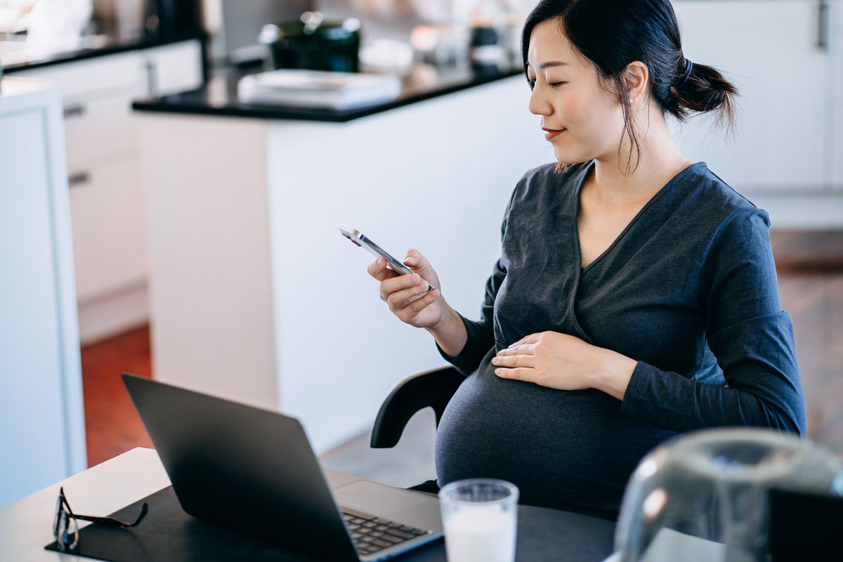 Professional young Asian pregnant businesswoman working from home, sitting at her desk in front of laptop and using smartphone. Hand gently touching her baby bump. Home office. Work from home. Mother working at workplace