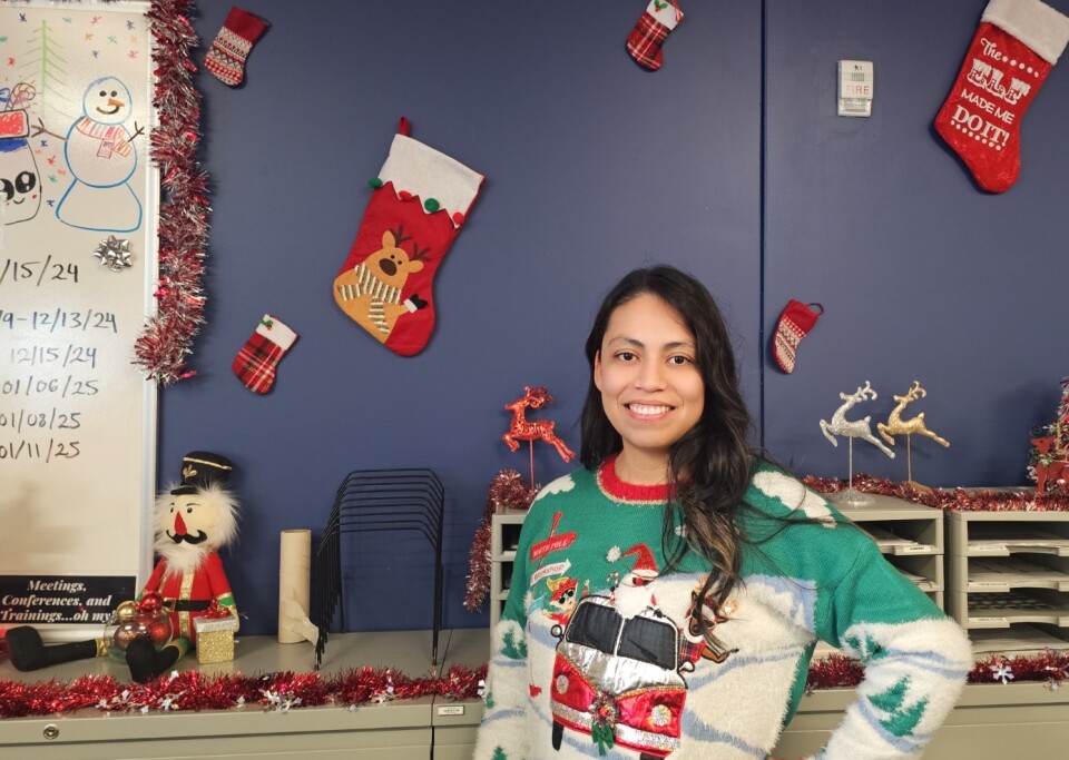 A woman with medium-light skin tone and long dark hair smiles for a photo. She wears a Christmas sweater with a Santa Claus and reindeer riding a red Volkswagen microbus. Behind her, there is a purple wall, as well as a whiteboard with a drawing of a snowman wearing a scarf. There are also other Christmas decorations in the background, including a stuffed nutcracker, reindeer, and stockings. 