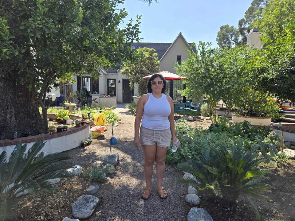 A woman with medium skin tone, wearing a white shirt and shorts, stands in a garden in front of a home with triangle peaks.