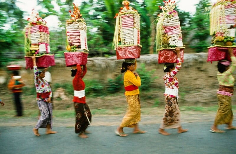A group of women in colorful traditional clothing walk barefoot in a line, each balancing an ornate offering basket on her head along a rural road with greenery in the background.