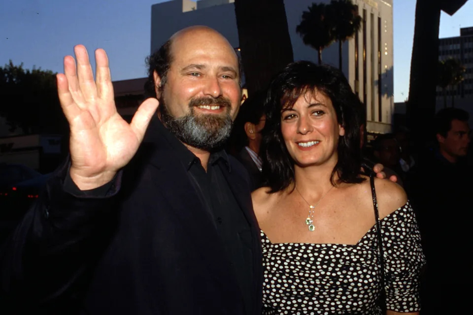 A man and woman smiling at an event, with the man waving at the camera. The woman wears a patterned off-shoulder dress