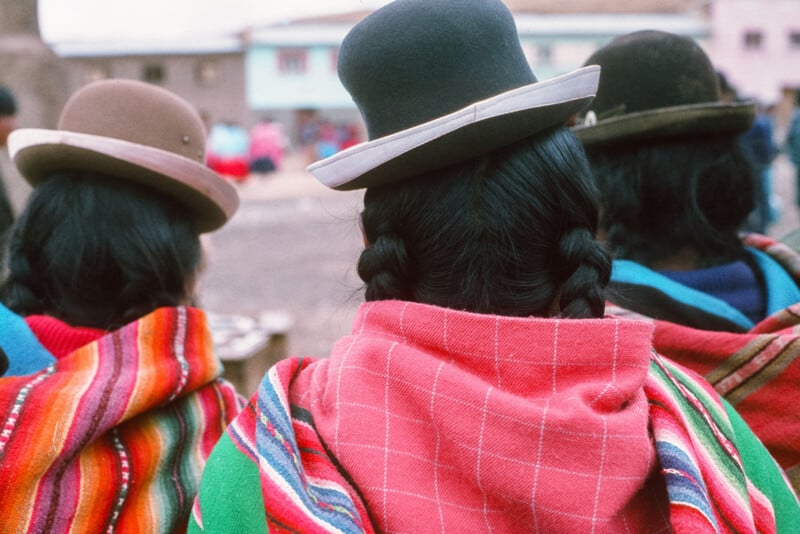 Three women seen from behind, wearing colorful traditional Andean shawls and bowler hats. Their dark hair is braided, and they appear to be outdoors in a village setting with blurred buildings in the background.