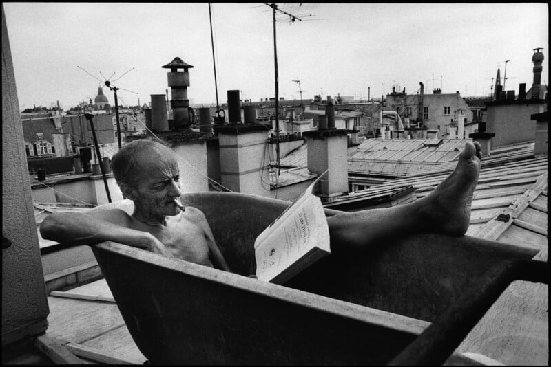 An older man relaxes in an old bathtub on a rooftop, reading a book with one leg propped up. The city skyline, chimneys, and antennas are visible in the background under a cloudy sky.