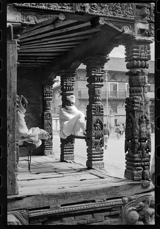 A black-and-white photo of two people sitting on the intricately carved wooden veranda of a traditional building, with one person facing away and a few people walking in the background.