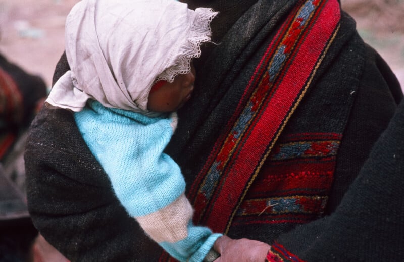 A small child wearing a light blue sweater and a white headscarf is carried on an adult’s back with a colorful woven textile, showing only the child’s profile and part of the adult.