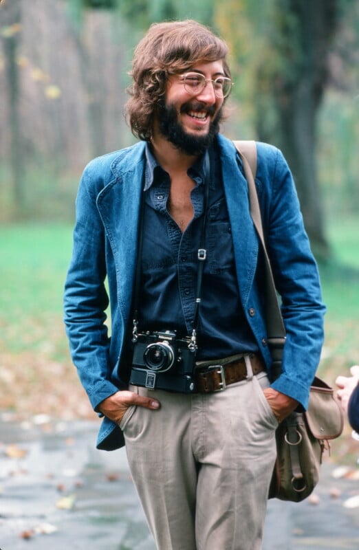 A smiling man with glasses, a beard, and long hair wears a blue jacket, navy shirt, and beige pants. He has a camera around his neck and a messenger bag over his shoulder, standing outdoors on a path with greenery in the background.