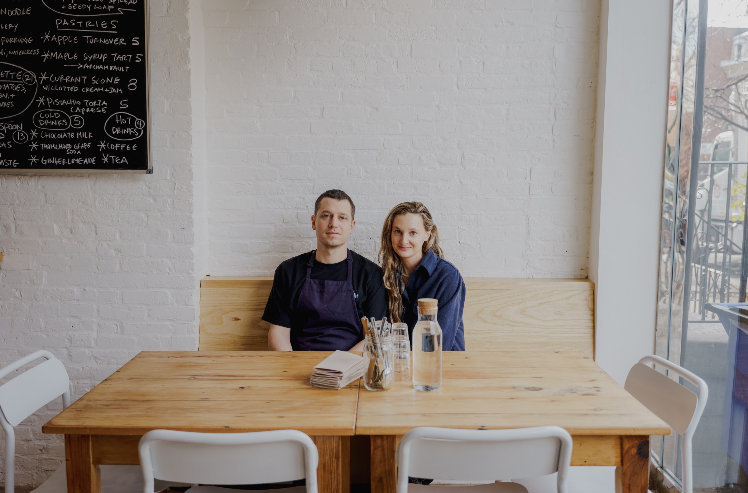 A man and a woman seated at a wooden table in front of a white wall.