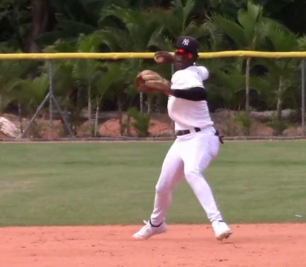 Wandy Asigen, a baseball player, in a white uniform, black New York Yankees hat, and red sunglasses, throwing a ball while wearing a mitt on a baseball field.