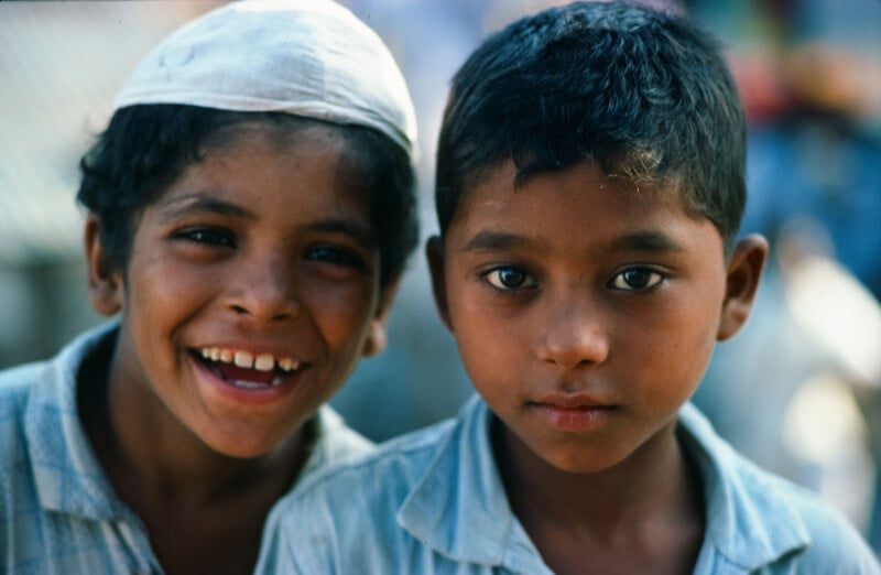 Two young boys stand close together, one smiling widely and the other with a neutral expression. Both wear light-colored shirts, and one boy wears a white cap. The background is blurred and outdoors.