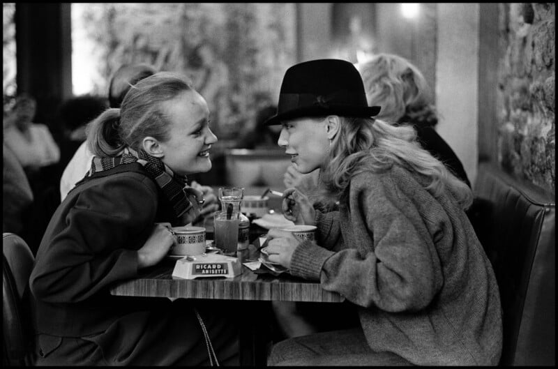 Two women sit closely at a café table, smiling and leaning toward each other in conversation. They both have drinks and bowls, and one wears a scarf while the other wears a hat and loose sweater. Other patrons are in the background.