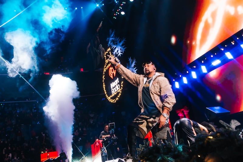 Sean Paul in brown jacket, black shirt, and camoflauge shorts performing on stage.