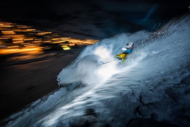A skier in bright gear carves through snow at night, illuminated by headlamps and city lights in the blurred background, creating a dynamic and energetic scene.