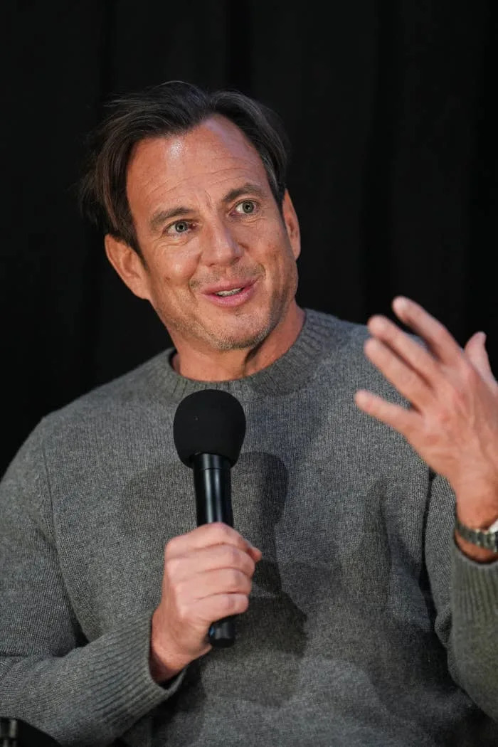 Person smiling and speaking into a microphone, wearing a casual long-sleeve sweater, in front of a black background