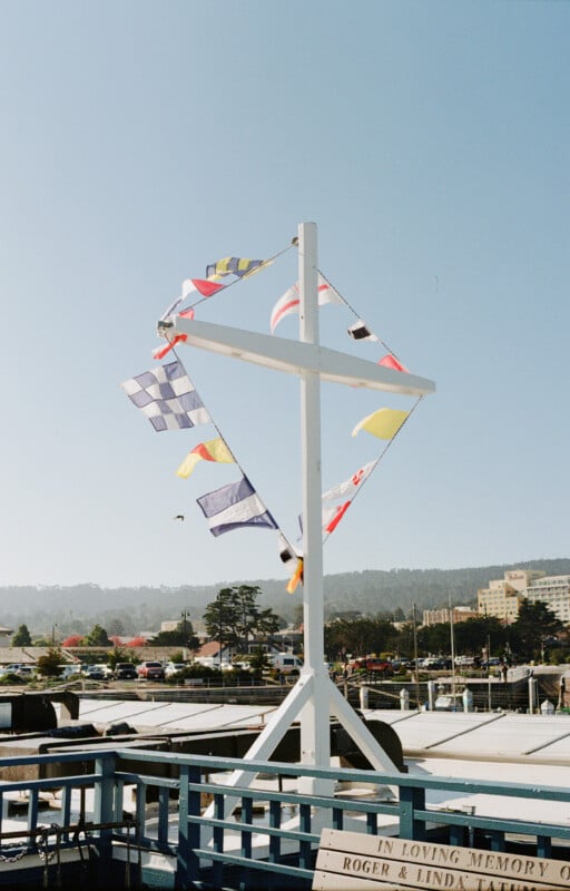 A white pole displays colorful nautical flags arranged in a diamond shape above a marina, with boats, trees, and distant buildings visible under a clear blue sky.
