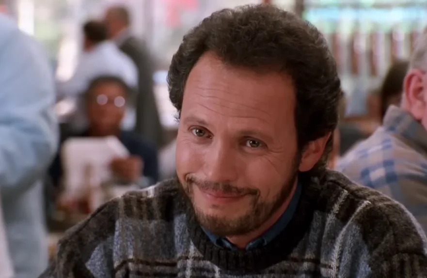 A person with curly hair and a beard smiles warmly while seated in a casual setting, wearing a patterned sweater