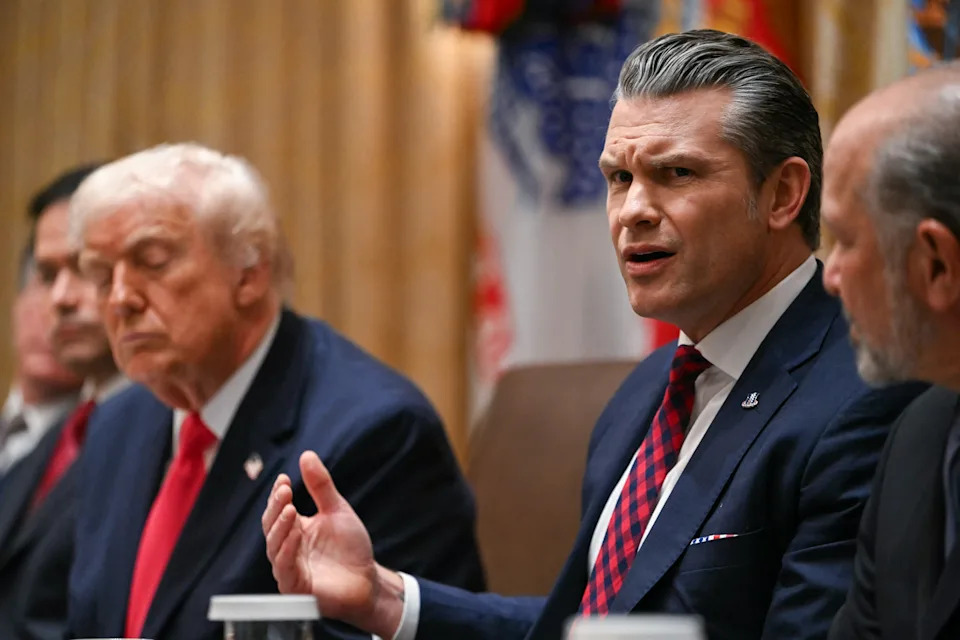 Men sitting at a roundtable discussion, one man speaking animatedly, others listening intently. Flag images in the background
