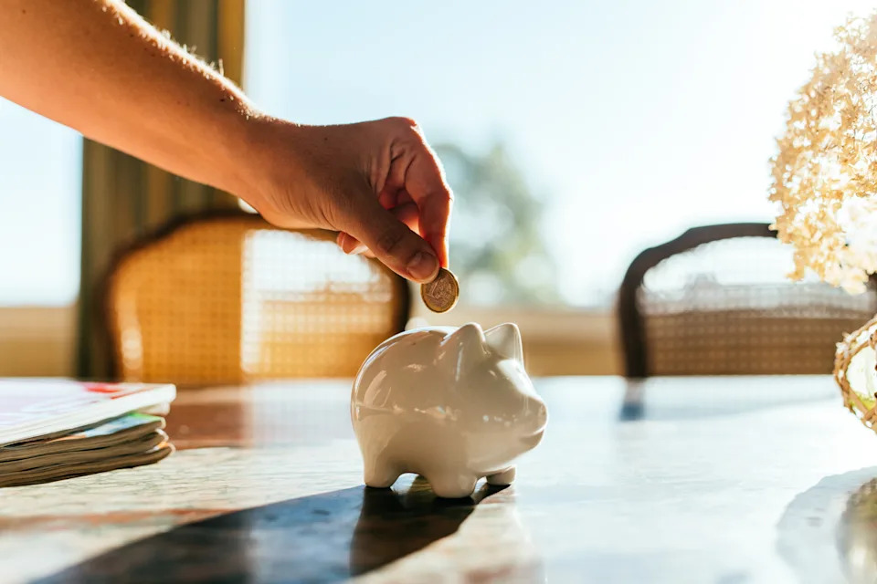 Hand placing coin into white piggy bank on a table, symbolizing saving money or financial planning. Bright sunlight streams in from a window