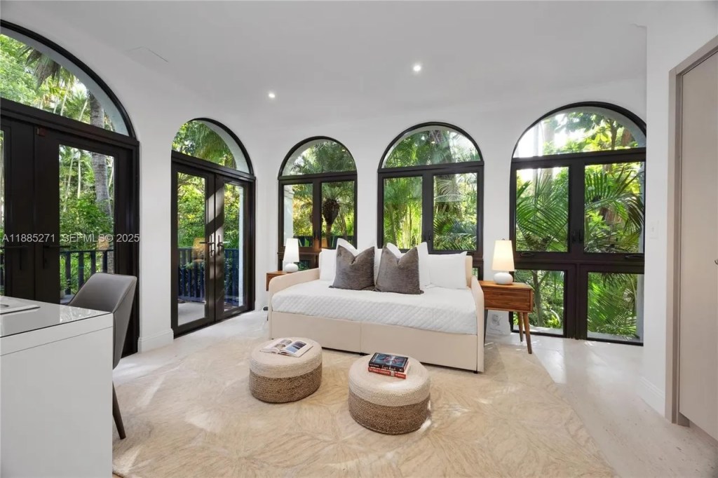 a spacious bedroom is shown with five floor-to-ceiling windows letting in sunlight and images of the greenery outside