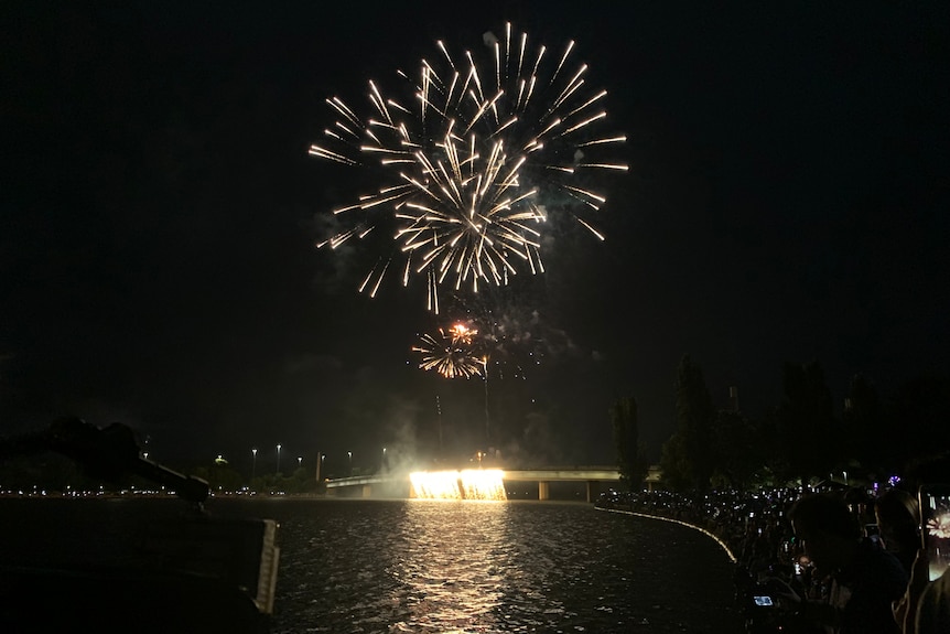 A wide shot at night of fireworks above a bridge on Lake Burley Griffin. Some fireworks flowing off the side of the bridge.