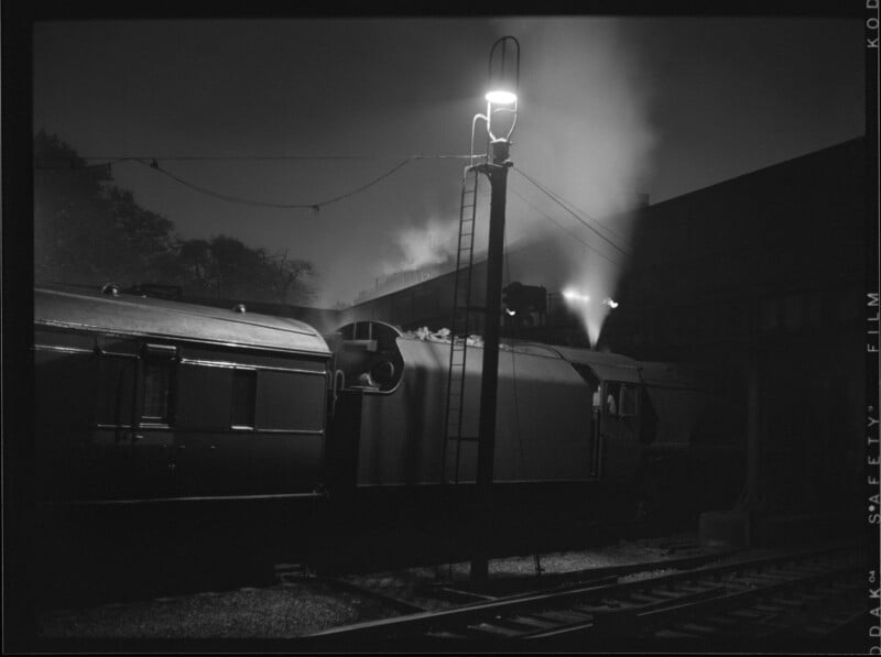A steam train at night emits smoke under a tall lamppost by the tracks; the scene is dimly lit, creating a dramatic silhouette against the night sky.