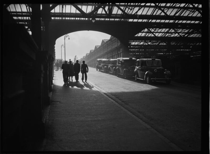 A group of people walk in shadow under a large metal bridge, while vintage cars are parked along the street beside a railway station, with sunlight streaming through the glass roof above.