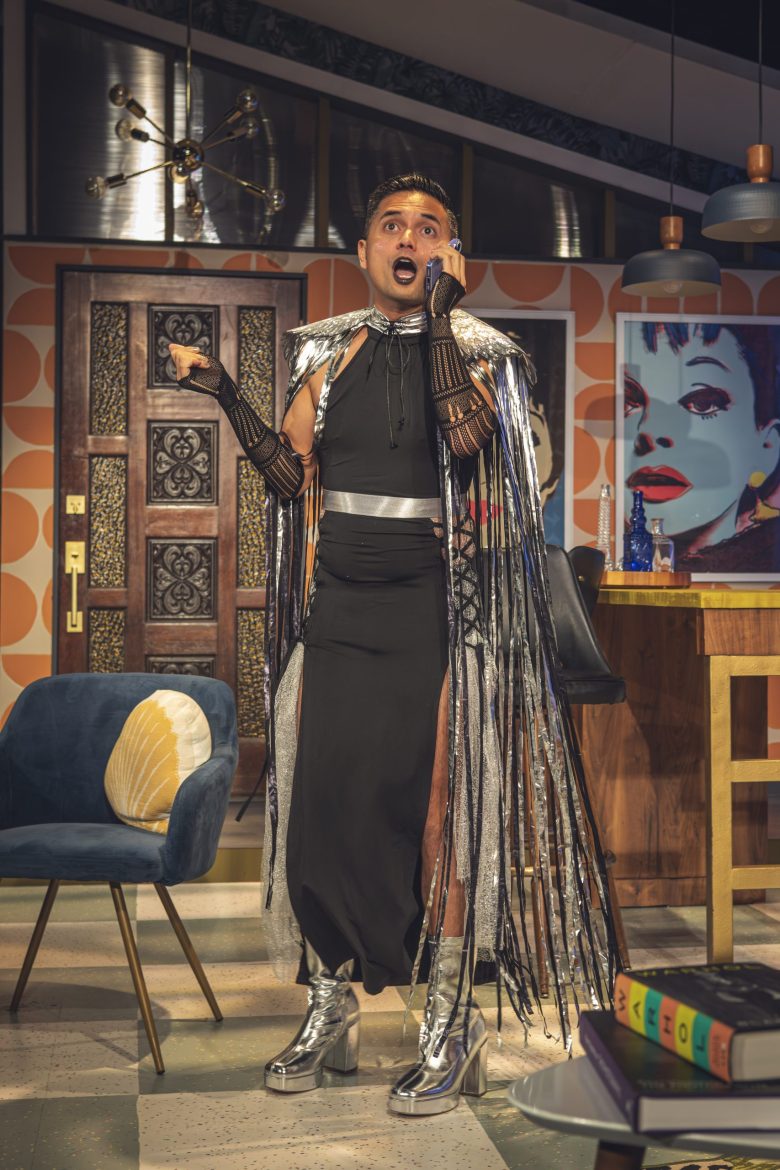 An Asian man wears a black dress with split sides and a feathery cape. 