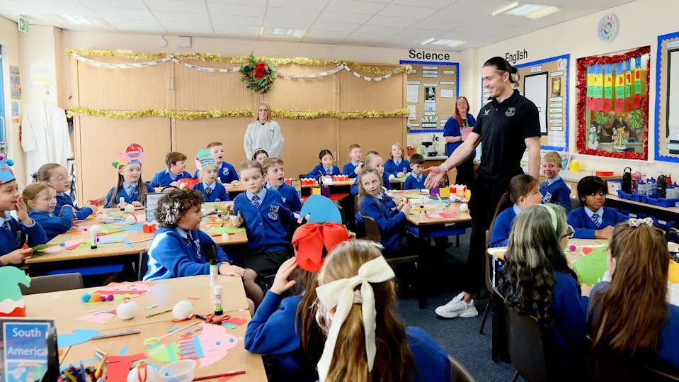 Jack Grealish stand and smailes at the front of a classroom with about 20 children. The children are spread around five big tables with various Christmas cutouts in front of them. The children looked surpised to see him as he moves to shake hands with Georgie at his table. Tinsel and school artwork decorate the walls around the room.