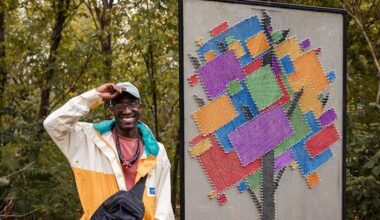 A person in outdoor clothing stands in a wooded setting with a standing public sculpture resembling a quilted tree in block colors of purple, red, green, yellow, and blue.